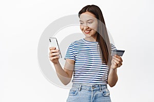 Young 20s woman paying online, shopping in app, confirm purchase with credit card info, looking at smartphone screen and