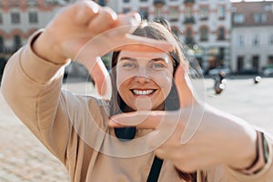 Young 30s female looking through a frame made from her fingers outdoors in the city. Happy cheerful young woman in