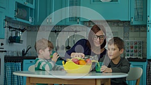 Young 30s mother with two kids drinking fruit juice sitting at the table with fruits on blue kitchen. Happy family enjoy