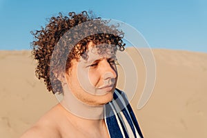 Young 32 years old Caucasian man with curly hair with striped towel on sandy beach on sand dune. Close-up summer headshot