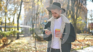 Young absorbed man messaging online in smartphone app walking in sunshine on autumn park alley. Portrait of concentrated