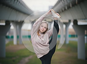 Young acrobat girl in beige hoodie performs stretching standing on one leg against the background of the bridge