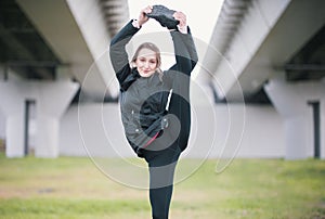Young acrobat girl in black jacket performs stretching standing on one leg against the background of the bridge