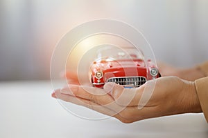 Young active business woman hands in casual dress sitting while holding and protecting car model or automobile on white table.