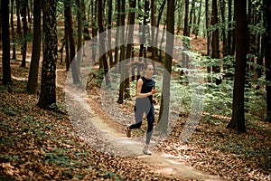 Young active fit woman running on narrow forest path between tall leafy trees