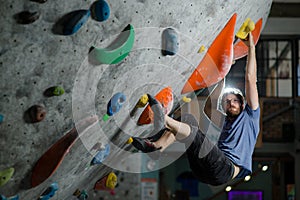 Young active sport caucasian american bearded man in glasses and t-shirt climbing on wall with color holds during bouldering