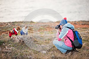 Young active woman in wool hat and track suit and backpack taking pictures of two little chihuahua pet dogs using camera outdoors