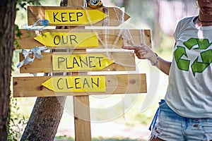 Young activist showing to wooden board saying, Keep our planet clean. Volunteers in nature picking up trash. Environmentalism,