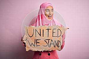 Young activist woman wearing pink muslim hijab holding banner with united we stand message pointing and showing with thumb up to