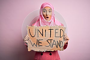 Young activist woman wearing pink muslim hijab holding banner with united we stand message scared in shock with a surprise face,