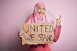 Young activist woman wearing pink muslim hijab holding banner with united we stand message surprised with an idea or question