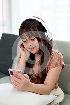 Young adorable Asian girl wearing headphones and using phone, enjoying listening to music on a sofa