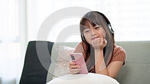 Young adorable Asian girl wearing headphones and using phone, enjoying listening to music on a sofa