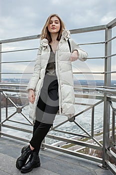 Young adorable blonde teen girl having fun on the observation deck with a view of cloudy spring sky, frozen river, sunny windy wea