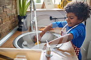 Young adorable boy with an apron, standing by the kitchen sink washing dishes, plates from lunch, helping with household chores