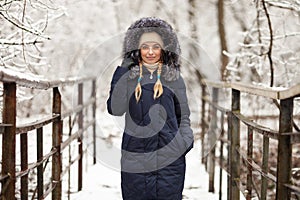 Young adorable woman wearing blue hooded coat enjoying strolling in winter forest outdoors. Nature cold season freshness concept.