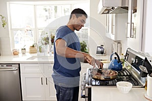 Young adult African American  man standing in the kitchen cooking on the hob, using a spatula and frying pan, side view, close up