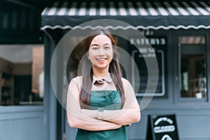 Young adult Asian business woman crossing her arms in front of cafe restaurant while smiling. Happy barista of Coffee