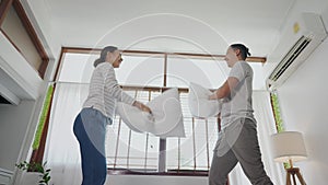 Young adult Asian couple playing pillow fight in bedroom interior scene. 30s mature husband and wife smiling and having