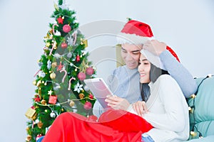 Young adult asian couple wearing santa claus hat using tablet computer on sofa in living room for online shopping with Christmas