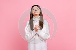 Young adult attractive brunette girl looking up with hands in prayer, sincere saying please, apology