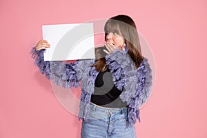 Young adult brown haired woman standing on pink background with whiteboard blank for sign on pink background in studio. Surpise,