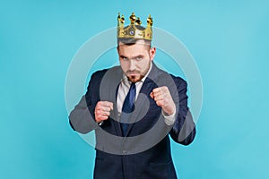 Young adult businessman wearing official style suit and golden crown, punching to camera, provoking fight or ready to self-defense