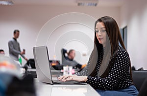 Young adult caucasian brunette woman sitting in an office environment looking at the laptop doing work. Multiple people in the