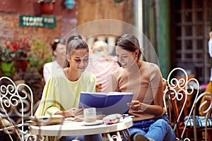 Young adult caucasian females reading notebook together, smiling, in the outdoor cafe