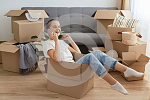 Young adult Caucasian woman wearing white t shirt and jeans sitting in a cardboard box and talking via mobile phone, boasting her