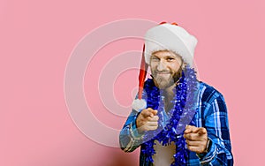A young adult cheerful guy 20-30s in a santa claus hat and new year tinsel makes a gesture with his index fingers forward