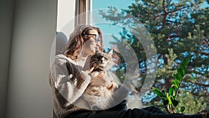 A young adult cheerful woman petting her cute cat while sitting on a windowsill