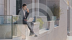 Young adult confident African American businessman using laptop outdoors holding smartphone, business negotiator on