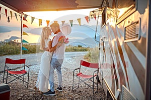 young adult couple hugging, holding each other in arms, in love, in front of a camper outdoor at the sunset and beautiful view