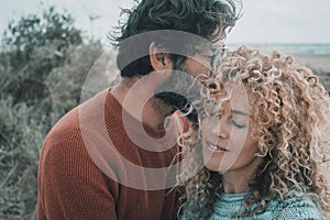 Young adult couple in love and tenderness outdoor leisure activity. Man kiss and whisper to a pretty woman with blonde long curly