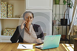 Young adult entrepreneur freelance black woman small business owner with laptop talking on mobile phone smiling in home