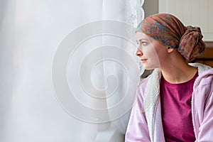 Young adult female cancer patient wearing headscarf and bathrobe sitting in the kitchen.