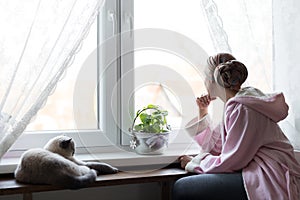 Young adult female cancer patient wearing headscarf and bathrobe sitting in the kitchen with her pet cat.