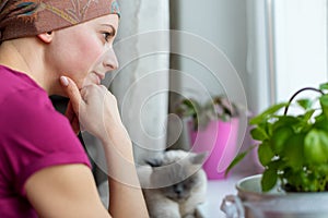 Young adult female cancer patient wearing headscarf and bathrobe sitting in the kitchen with her pet cat.