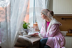 Young adult female cancer patient wearing headscarf and bathrobe sitting in the kitchen with her pet cat.