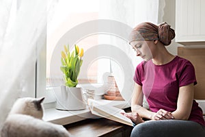 Young adult female cancer patient wearing headscarf sitting in the kitchen with her pet cat