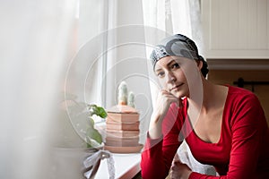 Young adult female cancer patient wearing headscarf sitting by the window in the kitchen.