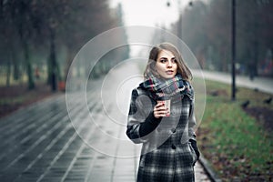 Young adult female with paper cup of coffee staying in city park on cloudy rainy day;