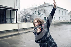 Young adult female with paper cup of coffee staying on the city street on cloudy rainy day and waving;