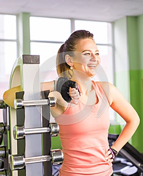 Young adult fit sporty girl posing near shelf with metal dumbbell in gym room space.