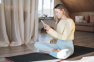 Young adult fitness blonde woman practice morning yoga using mobile phone near the window of home