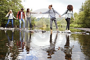 Young adult friends reaching to help each other cross a stream balancing on stones during a hike