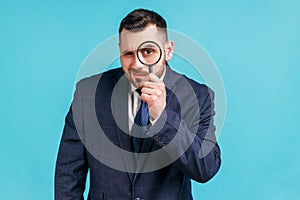 Young adult handsome businessman wearing dark official style suit holding magnifying glass and looking at camera with big zoom eye