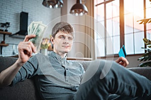 young adult handsome man choosing between cash and credit card on sofa at home