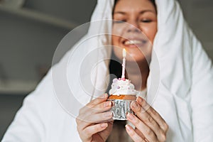 Young adult happy beautiful woman forty years plus size body positive in white blanket with festive cupcake with candle in hands